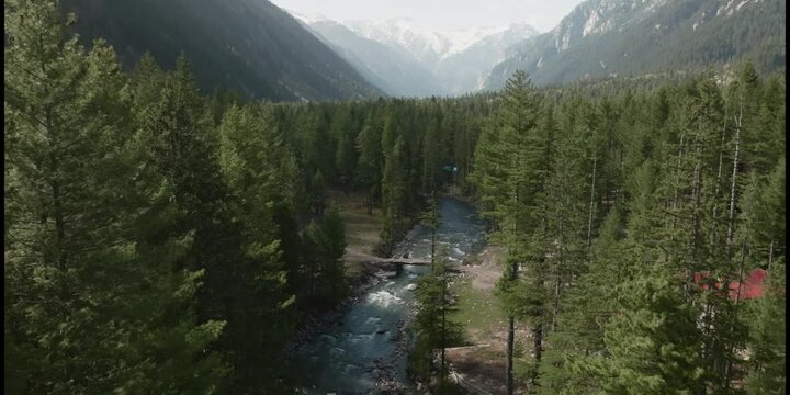 4 K Aerial shot, drone flights  in top of  river surrounded by close forest, camera tilts up a bit in the beginning and reveals snowed peaks in the back.  Kumrat, Pakistan, DJI Mavic 3
