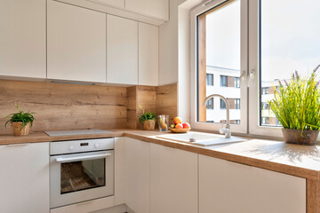 Modern kitchen with white furniture, wood on the wall, ceramic sink with tap, wooden counter and...