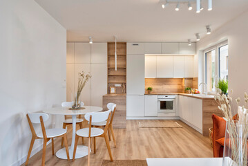 Kitchen interior with white furniture in scandinavian design and dining room with table and chairs. Interior of open space with wooden floor and window in new apartment in luxurious style.
