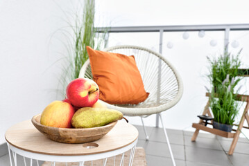 Stylish terrace with glass barrier and place for relax. Coffee table with bowl with fresh fruit, apple and pear and armchair with red pillow. Outdoor balcony with green plant. 