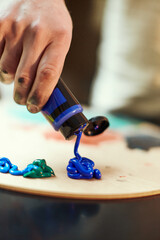 Artist squeezing blue acrylic paint on painting palette pressing tube in hand, preparing for creative work. Close-up