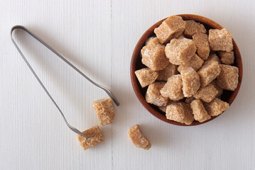 Cube-shaped cane sugar in wooden bowl on a white wooden table with silver sugar tongs.  High angle closeup, no people.