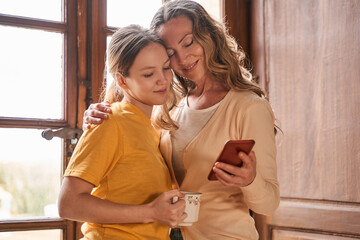 Woman holding smartphone and talking with her daughter while embracing at the kitchen