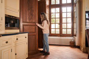 Calm adult woman standing in the kitchen and taking food from the big wooden cupboard