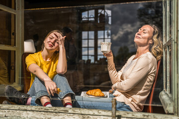 Smiling mother and teenage kid reposing near the window with mugs in hands