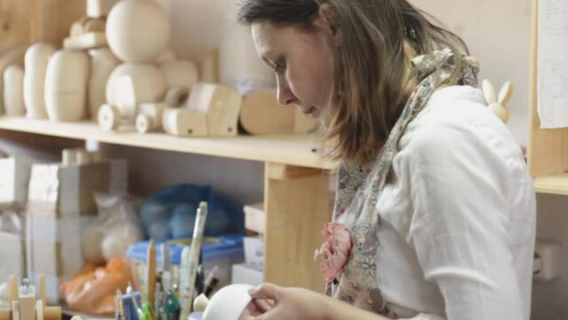 a female artist in the workshop carefully paints wooden toys with a brush. The craftsman is engaged in the manufacture of environmentally friendly toys.