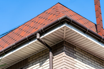 Brown roof of house covered with flexible bitumen motleysoft shingles under blue sky on sunny day. Cottage roof with metallic guttering system, guttering and drainage pipe exterior