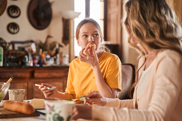 Caucasian teen girl eating and talking with her mother during the dinner at the kitchen