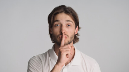 Young confident man  keeping fore finger on lips asking to make silence at camera isolated on gray...
