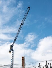 A construction tower crane froze against a bright blue summer sky on a sunny day against the backdrop of light white clouds.
