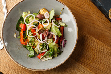 Greek salad with fresh vegetables: tomato, cucumber, red bel pepper, lettuce, onion, olives and cheese. Close-up on a white round plate on a wooden background. Salad menu with ingredients