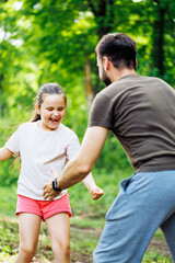 Portrait of family playing game of catch in park forest around trees, having fun. Little laughing girl daughter trying to run away from middle-aged bearded man father. Love, family, summer activities.