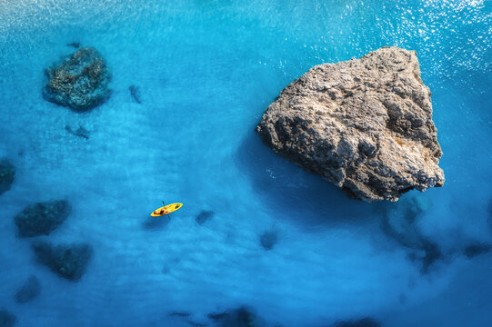 Aerial view of yellow kayak in blue sea at sunset in summer. Man on floating canoe in clear azure water, rocks, stones. Lefkada island, Greece. Tropical landscape. Sup board. Active travel. Top view