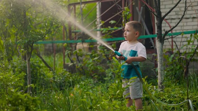 Cute toddler blond boy playing with garden sprinkler on summer hot day. Kid having fun on backyard spraying himself with a hosepipe. Happy childhood.