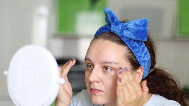 Curly woman in hairband applying and smearing moisturizing lotion or skincare cream for skin lifting. Circle and patting movements, home spa procedures treatment. Looking in mirror, facial massage. 