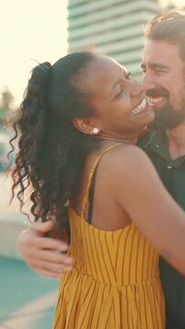 VERTICAL VIDEO: Close-up of young happy man and woman joyfully hugging when meeting on the embankment, man gives girl a bouquet of red roses. Closeup, joyful date of young interracial couple