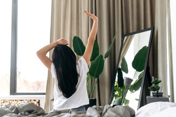 Happy asian brunette girl in a white t-shirt, is awakening and stretching arms after well sleep sitting in a cozy bed back to camera, starts her day with great mood, having a good night's sleep