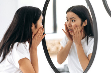Stunned worried Asian brunette girl, stands at home in front of a mirror, examines the skin of her...