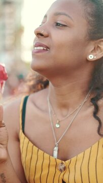 VERTICAL VIDEO: Closeup of smiling young woman with long curly hair on an urban city background with ice cream in her hands. Frontal close-up of happy girl testing ice cream on warm sunny day