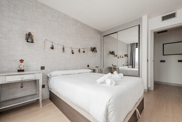 Fototapeta na wymiar bedroom with double bed with white duvet and built-in wardrobe with mirrored doors in one corner and decorative wallpaper on the wall
