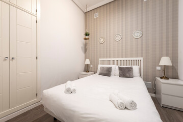 Fototapeta na wymiar bedroom with a double bed with a white duvet, decorative wallpaper on the wall, white bedside tables with twin lamps and a built-in wardrobe with white doors