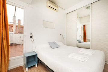 Fototapeta na wymiar Bedroom with a double bed with a white bedspread, a built-in wardrobe with Venetian-style white wooden sliding doors, another with a mirror, blue bedside tables and access to a terrace