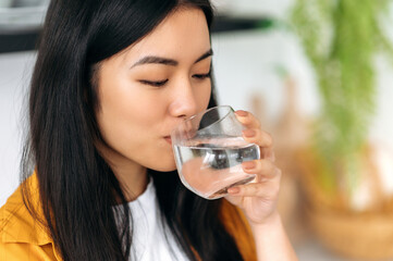 Leading a healthy lifestyle. Close-up of a happy Asian girl, drinks a glass of clean water with...