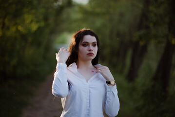 Sexy sensual brunette walks in a summer forest at sunset in a white blouse and jeans