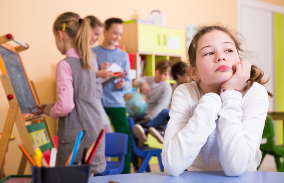 Upset girl sitting at table in schoolroom during break on background with other pupils