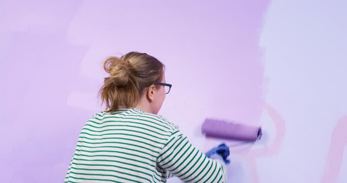 Young woman in glasses tries to paint over writing on wall in light purple using roller. Blonde lady in gloves does housework close backside close