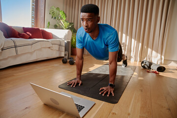 Young Black African American male working out at home holding a plank position about to do push ups on a yoga mat. Following an online class using his laptop.