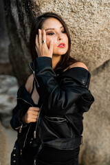 Fototapeta na wymiar Confident woman with red lips in trendy black leather jacket and sensual black dress posing on rocky background.