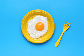 Fried egg on a bright yellow plate and a yellow fork on a blue background. A popular breakfast. Minimal concept. A popular breakfast.