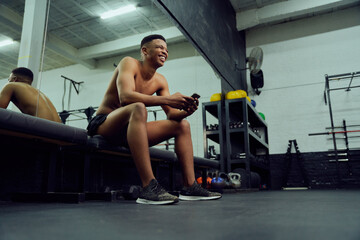 Fototapeta na wymiar Young African American male looking at his phone to reply to an online message. Happy, mixed race male smiling at his phone while sitting down in the gym. High quality photo 