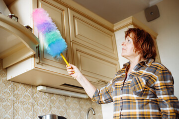 Portrait of elderly woman with dark hair standing in beige kitchen, wiping dust off cupboard with...
