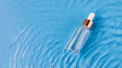 Glass bottle of anti aging serum and falling water drops on blue background. Facial serum mockup.
