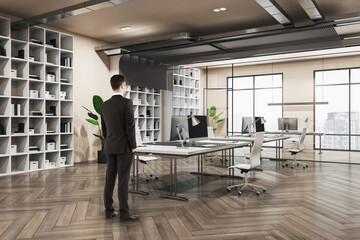 Handsome thoughtful young european businessman standing in wooden and concrete office interior with furnituire and window with city view. CEO, executive and consulting concept.