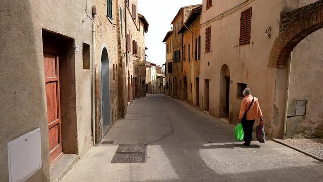 a lone older woman with shopping bags walks a lonely street in Siena Italy in morning sunshine