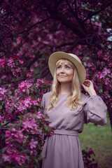 portrait young beautiful woman in a pink dress and hat stands by a blooming pink apple tree.