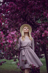 portrait young beautiful woman in a pink dress and hat stands by a blooming pink apple tree.