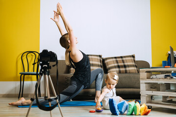 Young mother fitness instructor personal trainer record video near camera at home with baby daughter