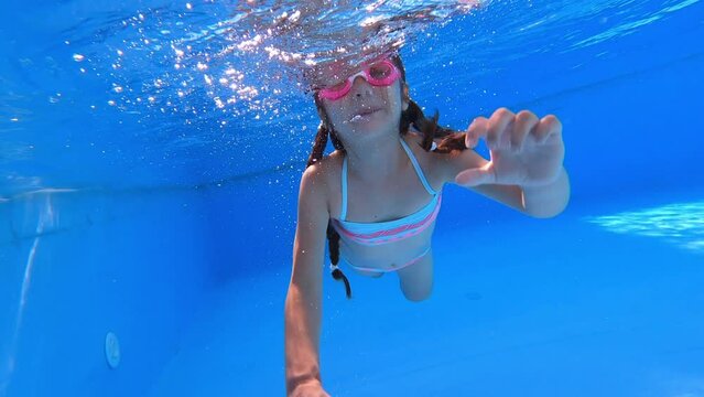 Little girl deftly swim underwater in pool and shows heart. Kid have blue bikini and pink glasses. Study swim. Sport good for healthy. Having fun in pool. Vacation and hobby concept