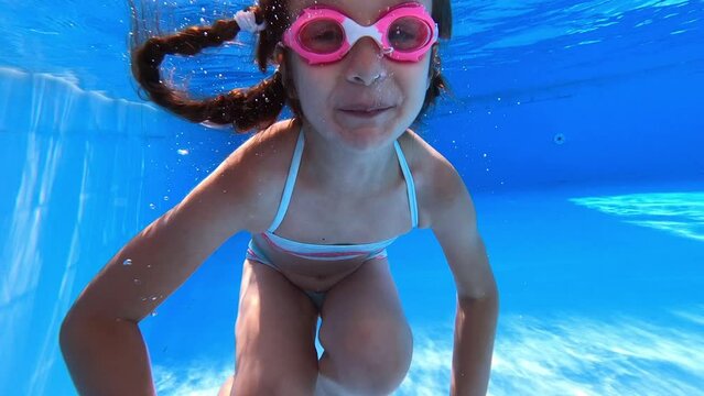 Little girl deftly swim underwater in pool. Child in blue bikini  and pink glasses jumping and holds breath. Sport good for healthy. Having fun in the pool. Vacation and hobby concept.