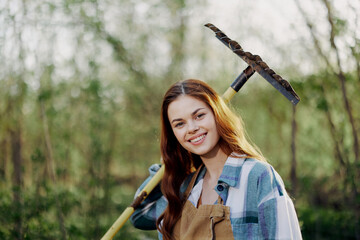 A woman smiling beautifully and looking at the camera, a farmer in work clothes and an apron working outdoors in nature and holding a rake to gather grass and forage for the animals in the garden