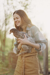 A happy young woman smiles at the camera and holds a young chicken that lays eggs for her farm in the sunlight