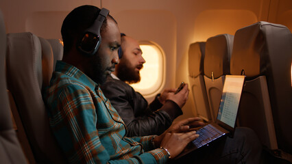 African american freelancer using laptop on flight, travelling on work trip during sunset in economy class. Working on computer and flying on airplane, aviation journey.
