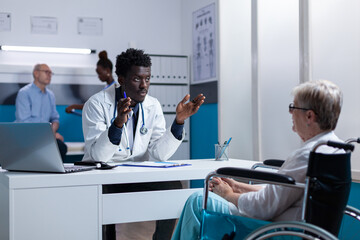 Physician expert explaining noninvasive procedure and treatment schedule to senior patient in wheelchair. Person with disability listening to hospital doctor talking about checkup appointment