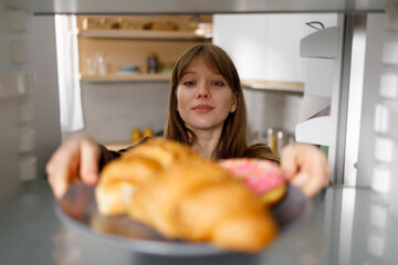 Young woman taking plate with croissants and donut from fridge