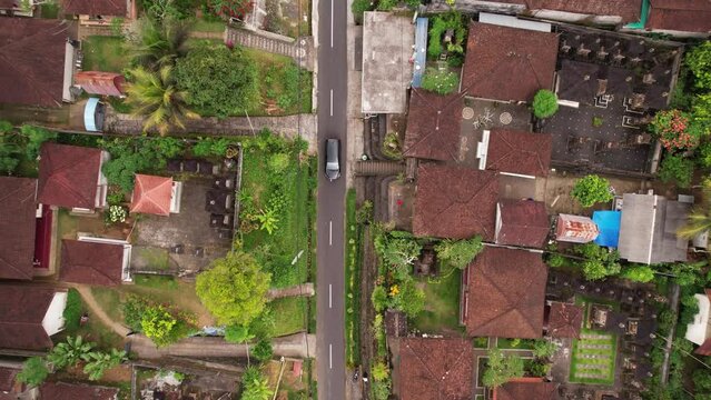 Panning aerial shot of small Balinese village, camera look straight down and follow one car riding at narrow street. Traditional houses seen along alley. Tegallalang area of Bali, near Ubud town