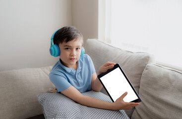 Kid wearing headphones holding tablet mockup, School boy using digital pad doing homework,Child sitting on sofa playing game online on internet with friend,Children with technology and Social media